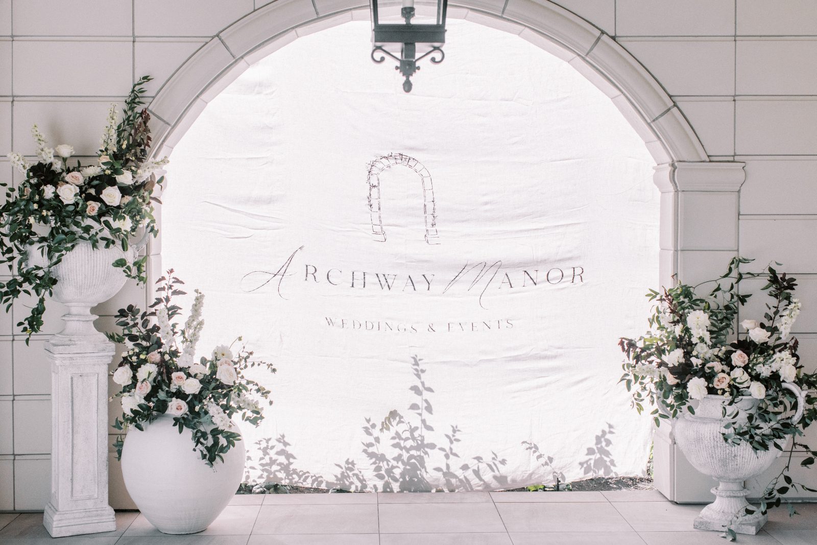 A pretty display for Archway Manor accented by large-scale Floral urns featuring creamy whites and blush flowers with lush greens and burgundy foliage.