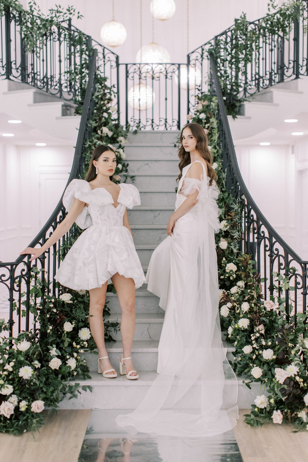 Two models pose on the stairs, on in a shorter gown with dramatic ruffles as sleeves, and the other with sheer billow cap sleeves that stream down behind her.