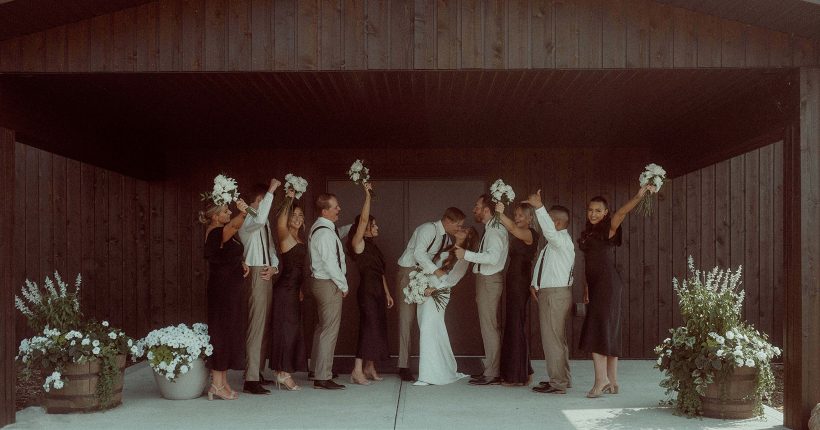 Allison and Taite share a kiss, while their wedding party surrounds them, cheering enthusiastically