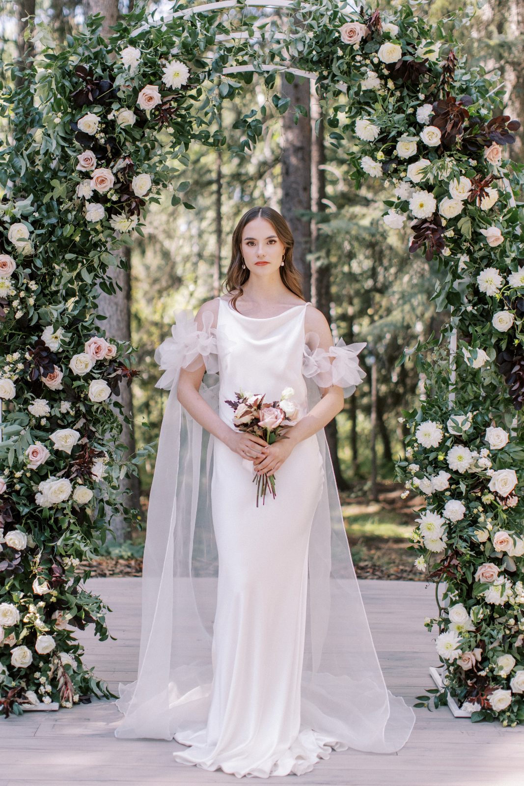 A model stands under an outdoor archway with tulle cap sleeves and cape-like train of tulle.