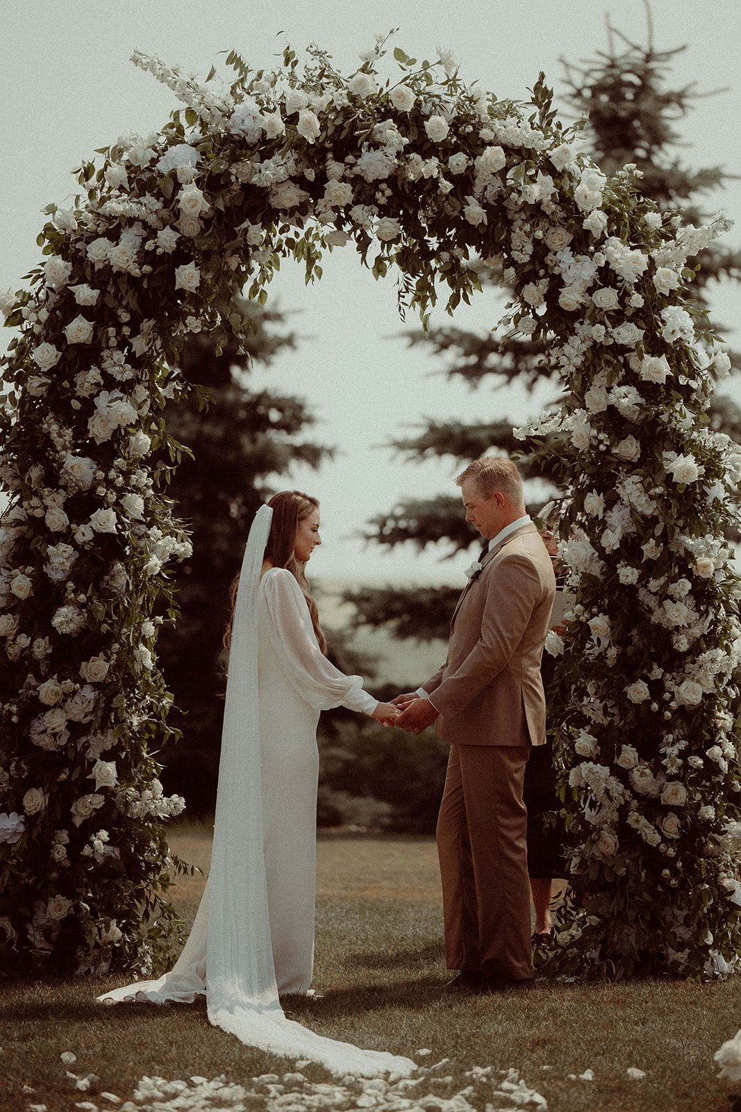 The beautiful Calyx Couple clasping hands at the end of the aisle underneath a dramatic, full arch of white and green.