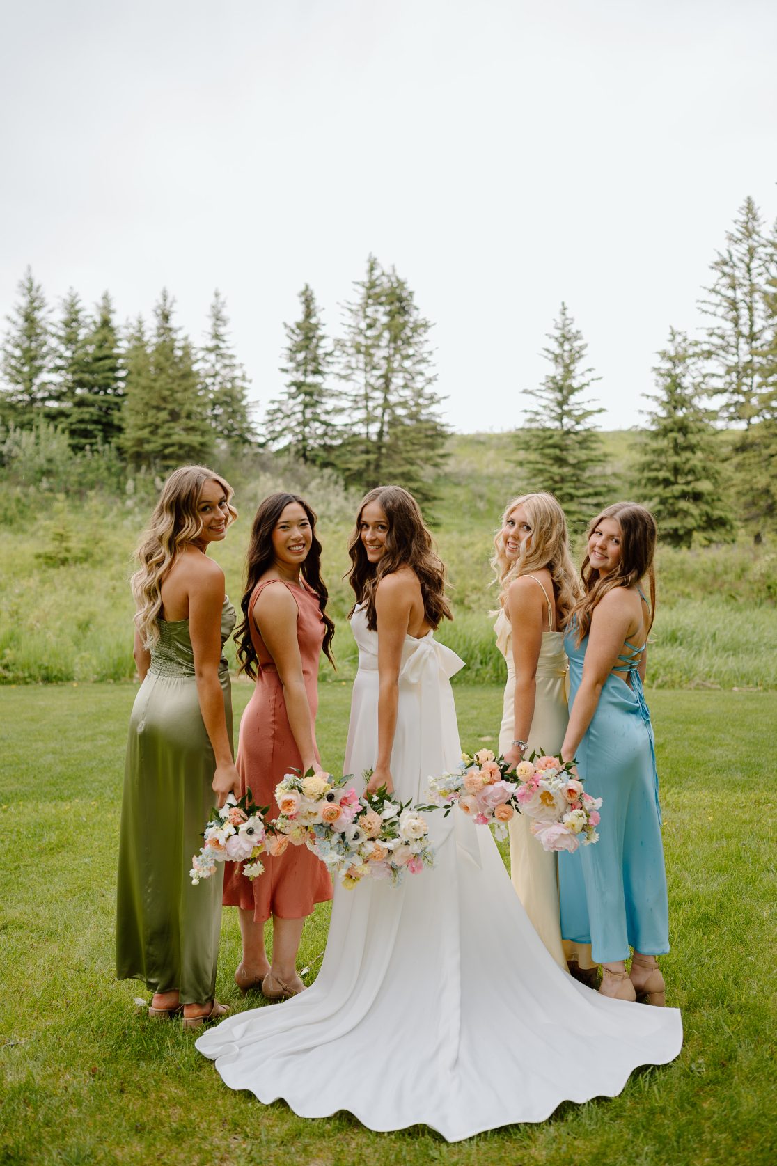 Mya stands with her dress fanned out behind her and her bridesmaids standing beside her, their bodies are faced away but they're looking over their shoulders smiling, with their bouquets by their sides.