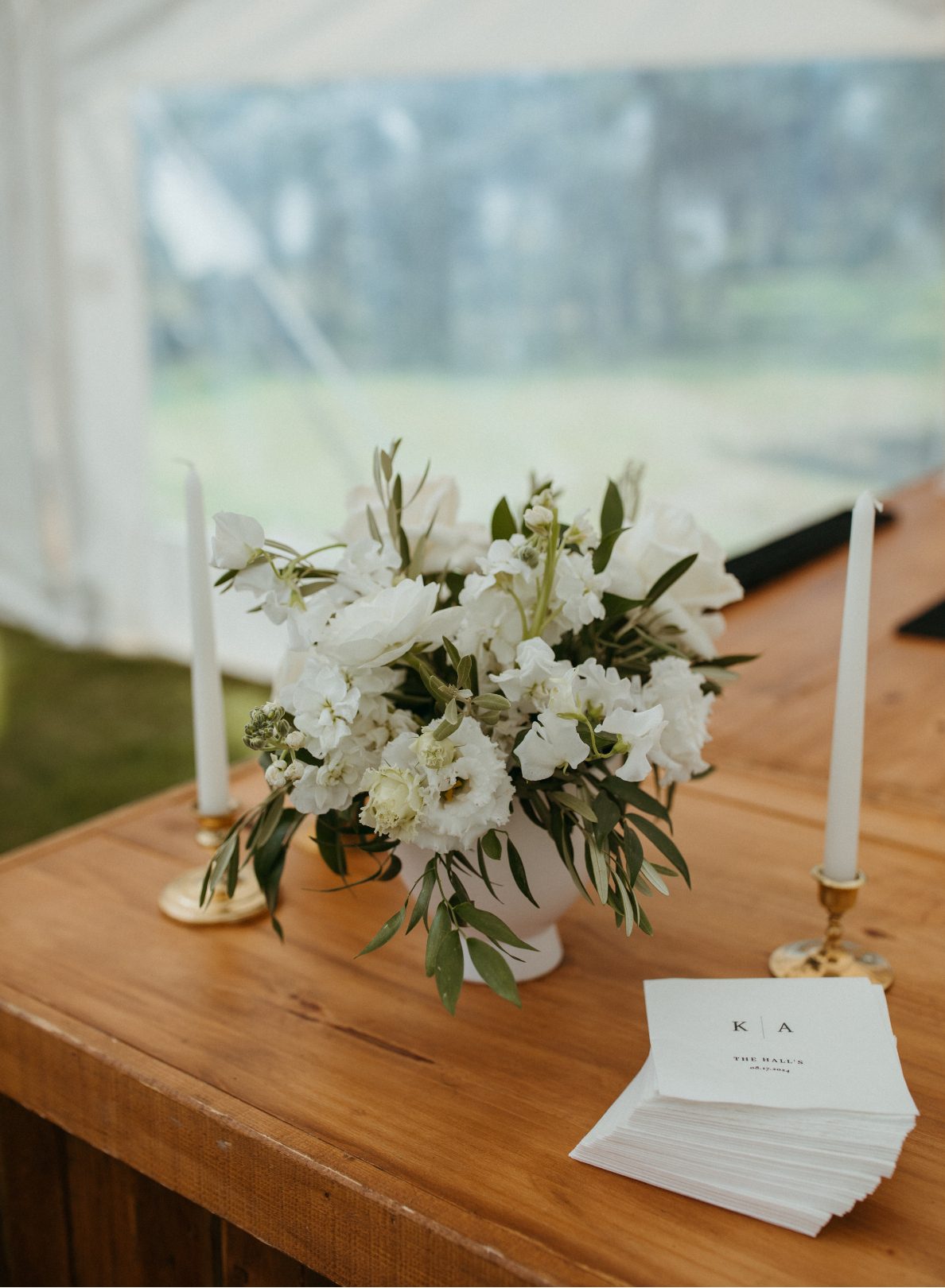 White floral arrangement in a white ceramic compote with olive greenery, sweet peas, lisianthus flanked by brass candlesticks with ivory taper candles on a wooden bar, next to personalized cocktail napkins reading "K+A, the Hall's."