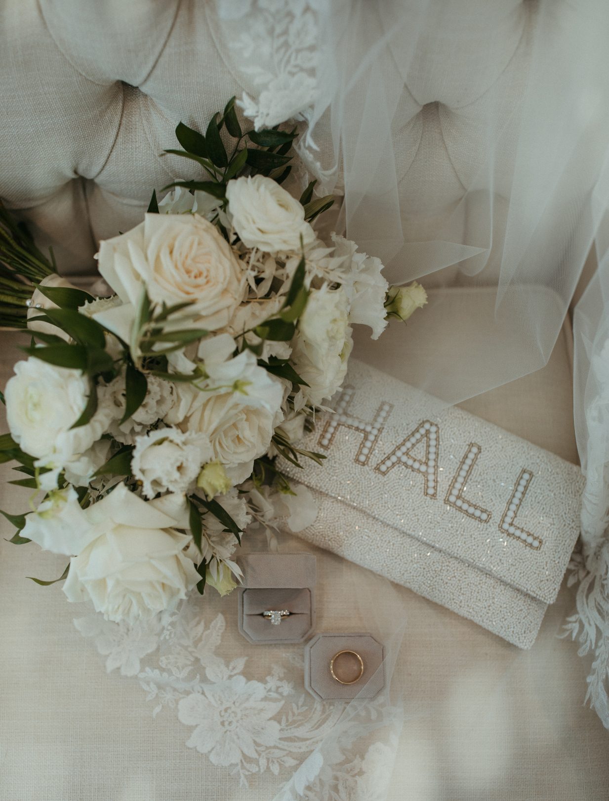 Flatlay of the bride's white bouquet, lace veil, sparlking personalized clutch reading "HALL," and wedding rings in velvet boxes