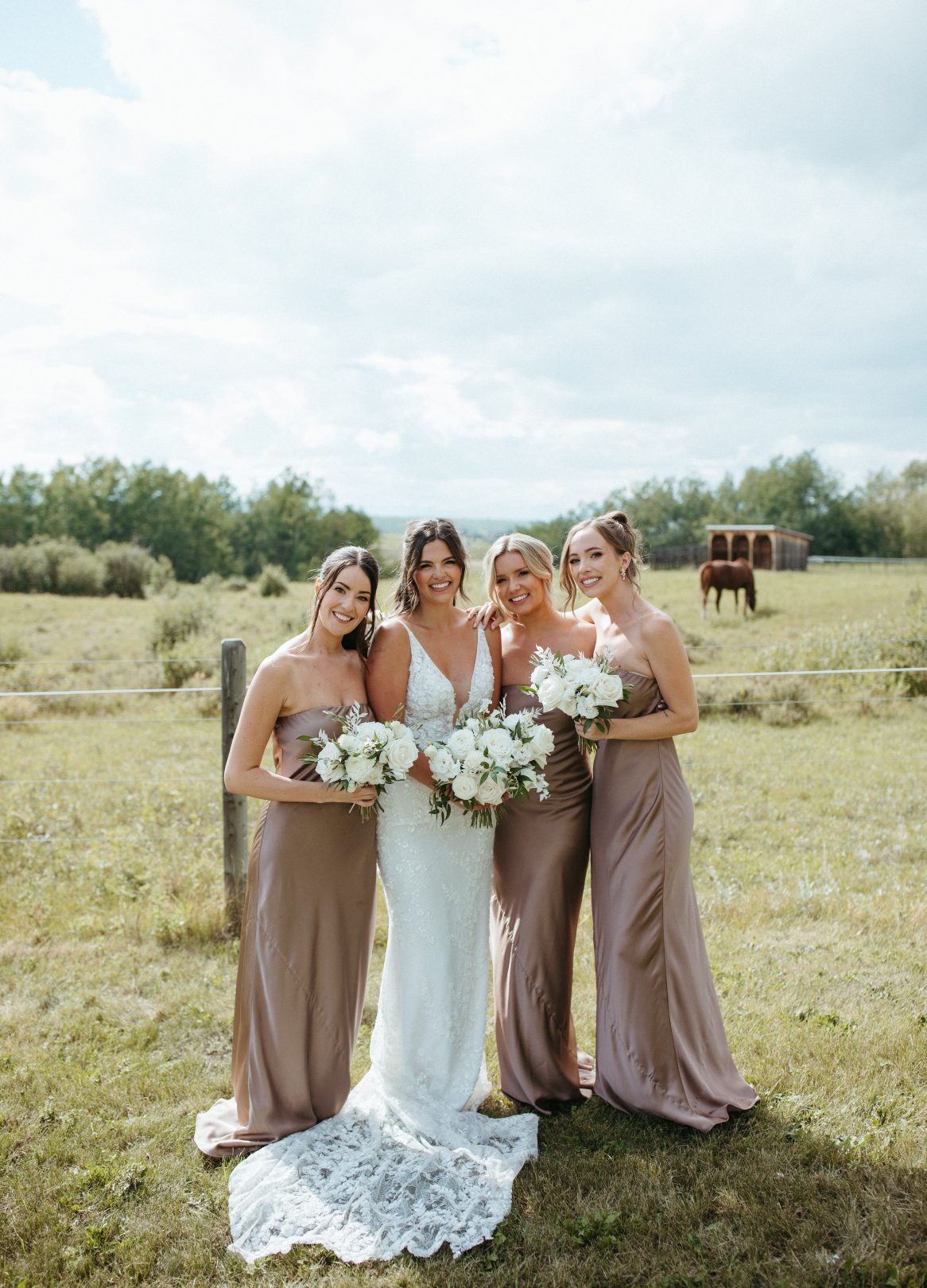 Bride and her three bridesmaids in silky taupe gowns, holding white bouquets, posing in a grassy field with horses in the background.