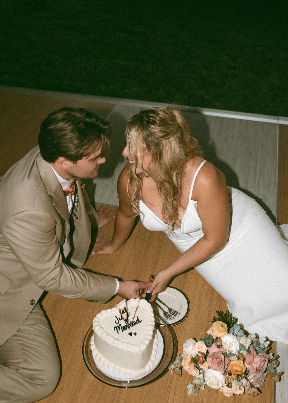 Katrina and Connor sitting on the dance floor looking at each other and laughing as they cut the heart shaped cake.