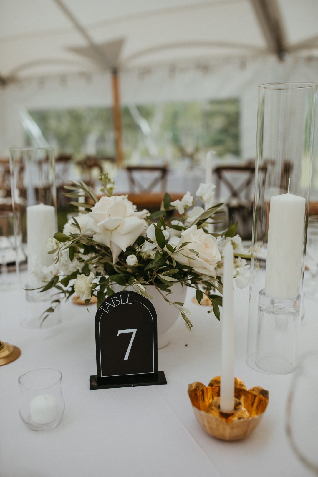 Elegant wedding centrepiece with white roses, lisianthus, olive greens and ruscus arranged in a white bowl, surrounded by white taper candles in assorted brass and glass holders, set on a white linen table with black and white arched table numbers.