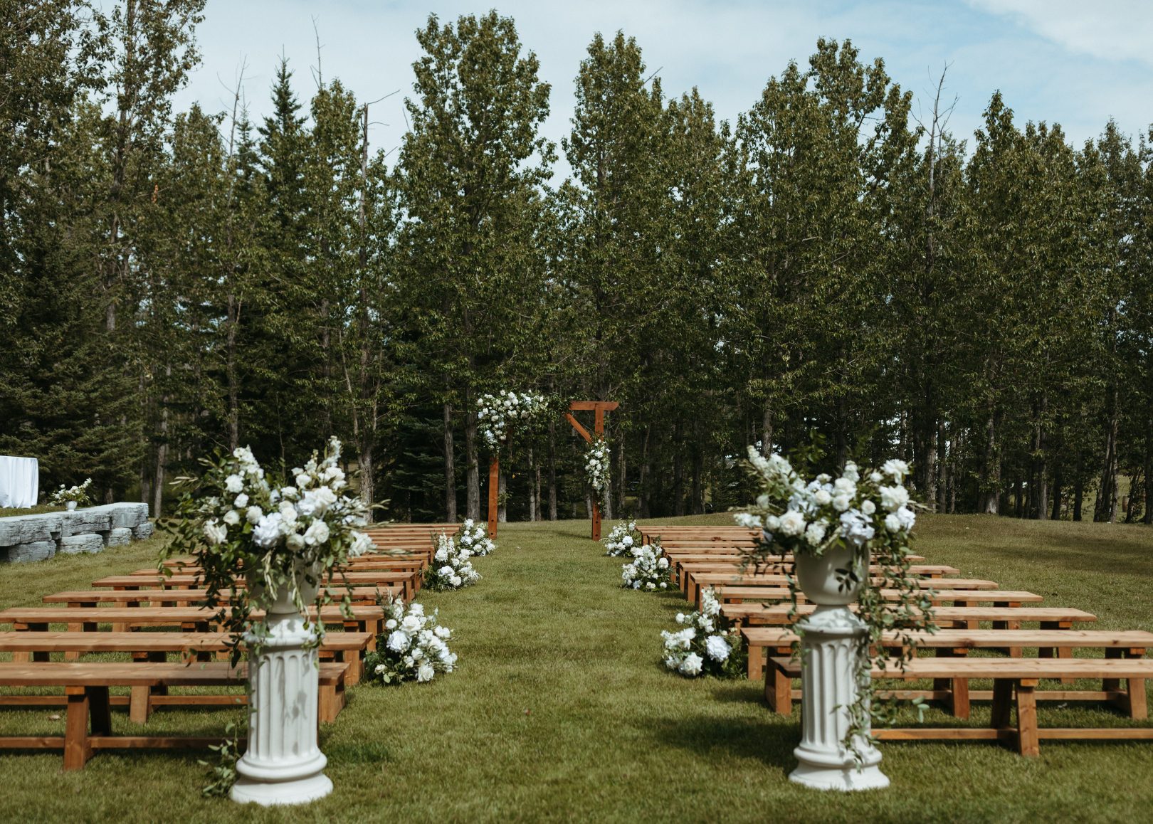 Wooden benches line a lush green aisle, flanked by grand floral arrangements in white and ivory with a background of towering trees and a beautiful floral arch at the end of the aisle.