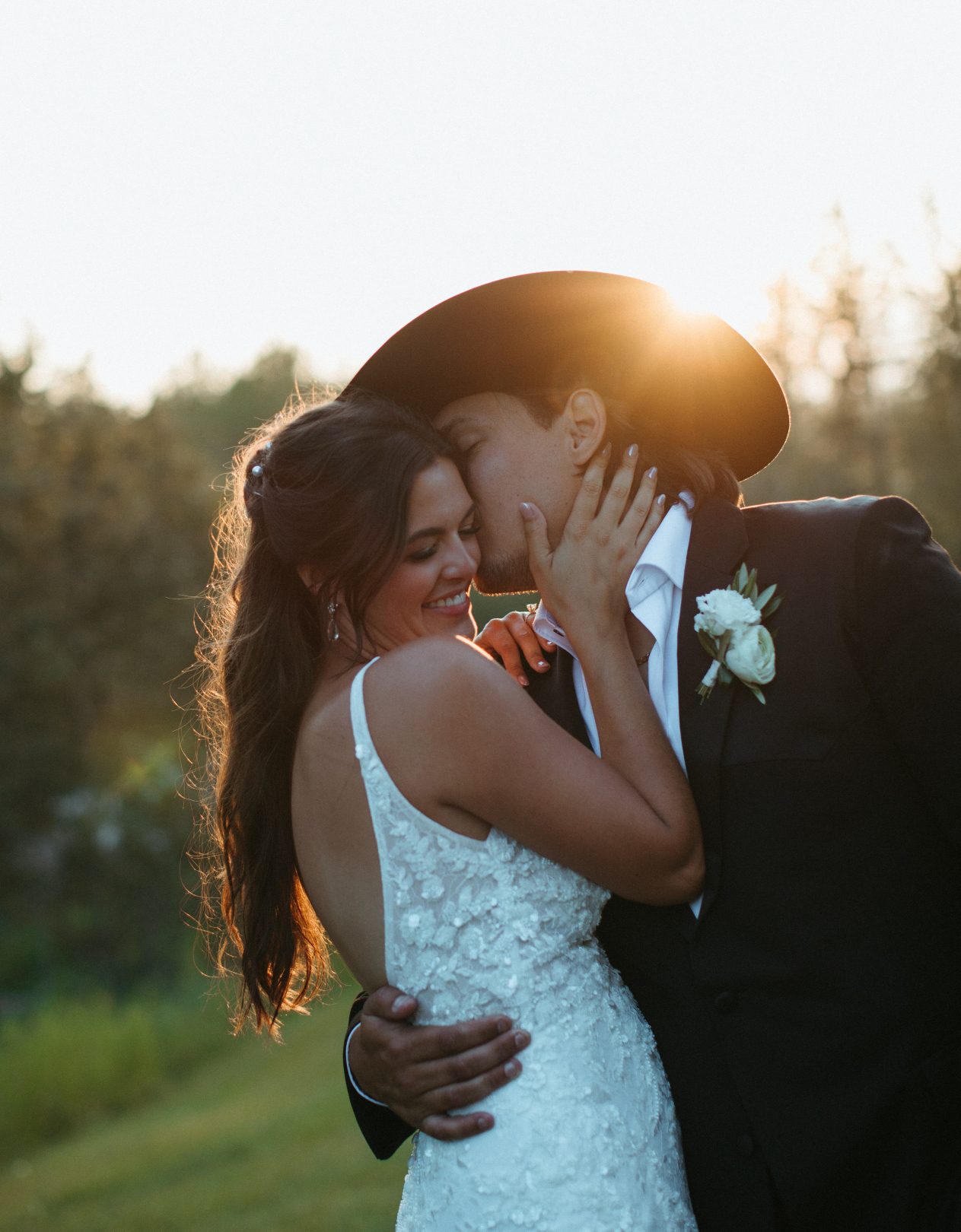 Groom in a black cowboy hat kisses bride on her temple at golden hour, with sunlight glowing behind them.