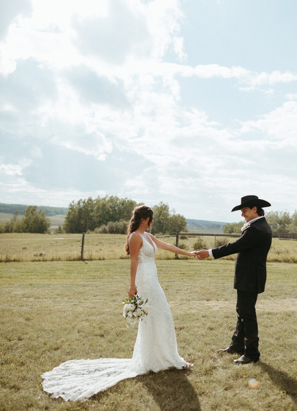 Bride in a lace dress and groom in a cowboy hat hold hands in a field, her train displayed behind her, with big blue skies overhead.