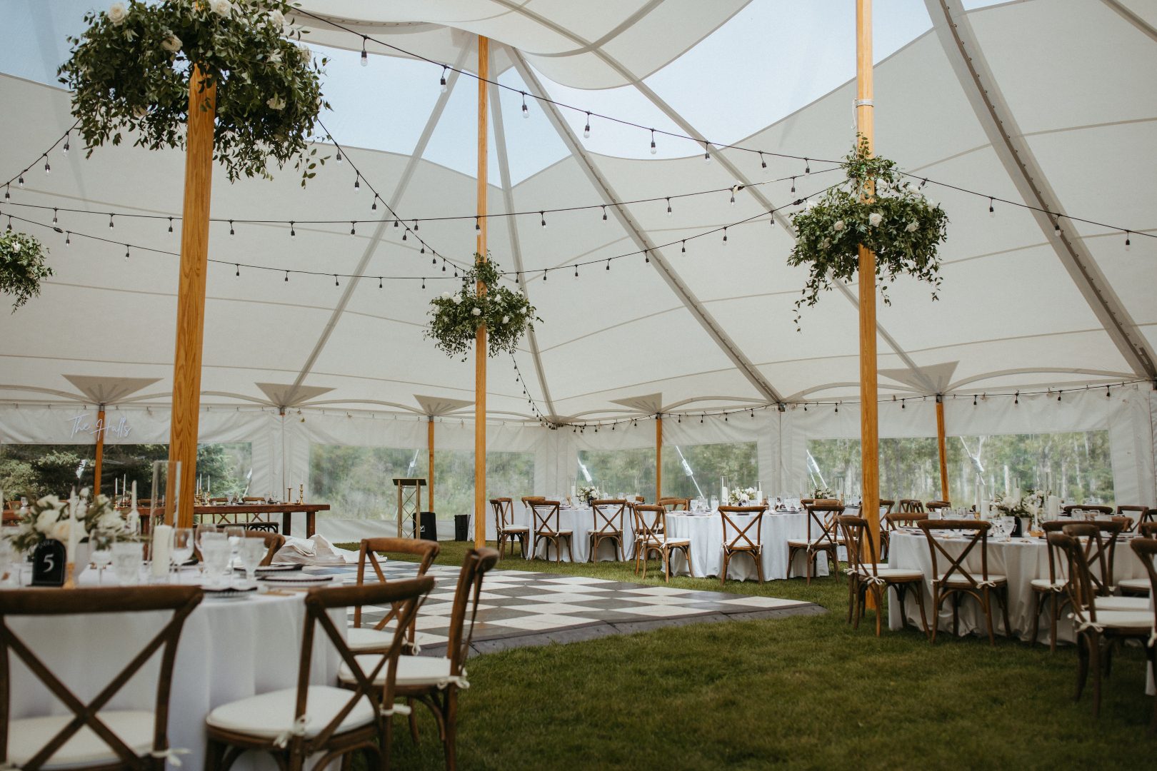 Spacious tent reception set up with round tables, cross back charis, twinkle lights, and lush greenery chandeliers suspended from tall wooden poles above a checkered dance floor.