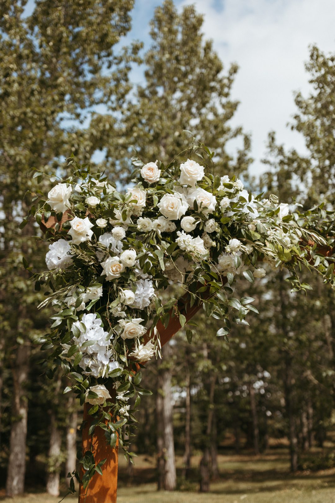 A romantic floral arch detail featuring ivory roses, white hydrangea, ranunculus and ruscus, beautifully arranged on a rustic wooden frame.