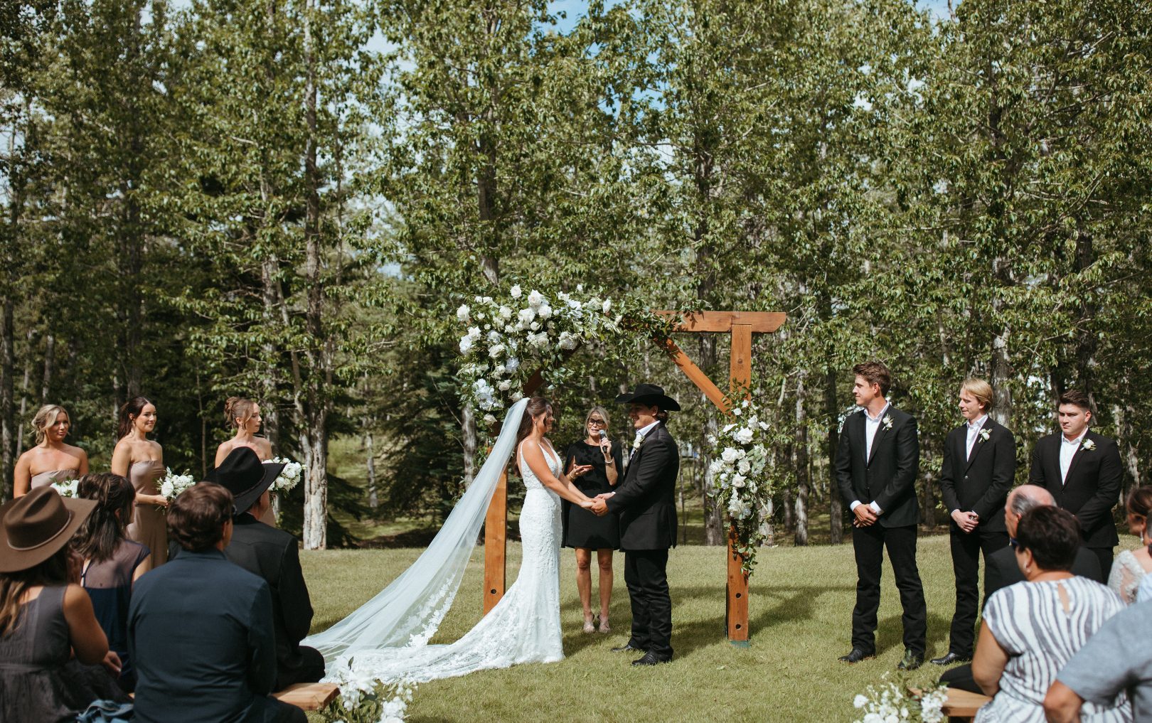 The bride and groom stand facing each other, hlding hands beneath a wooden arch lushly adorned with white flowers and greenery while exchanging vows surrounded by their bridal party and loved ones.