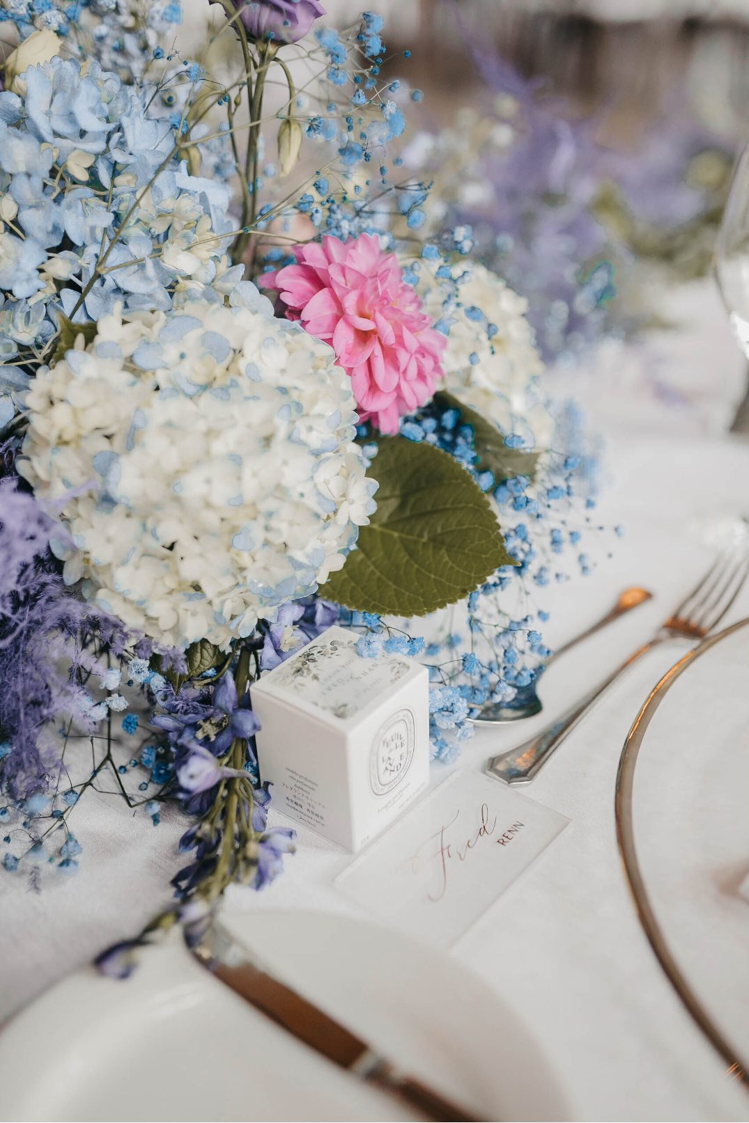 A lush arrangement of ydrangea, pink dahlia and baby's breath next to a candle favor and name card for Fred.
