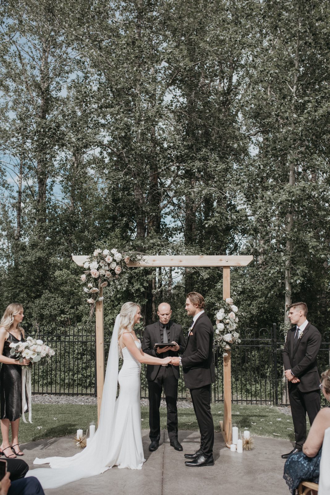 The bride and groom hold hands during their outdoor ceremony beneath a wooden arch adorned with blush and ivory rose surrounded by soft greenery.