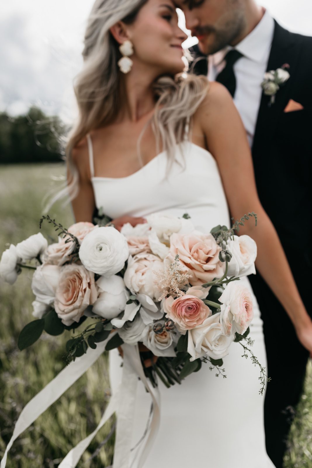 A soft-focus image of the bride and groom leaning in close, with a detailed view of the bride's lush bouquet featuring blush roses, white ranunculus, astilbe, eucalyptus and trailing silk white ribbon.