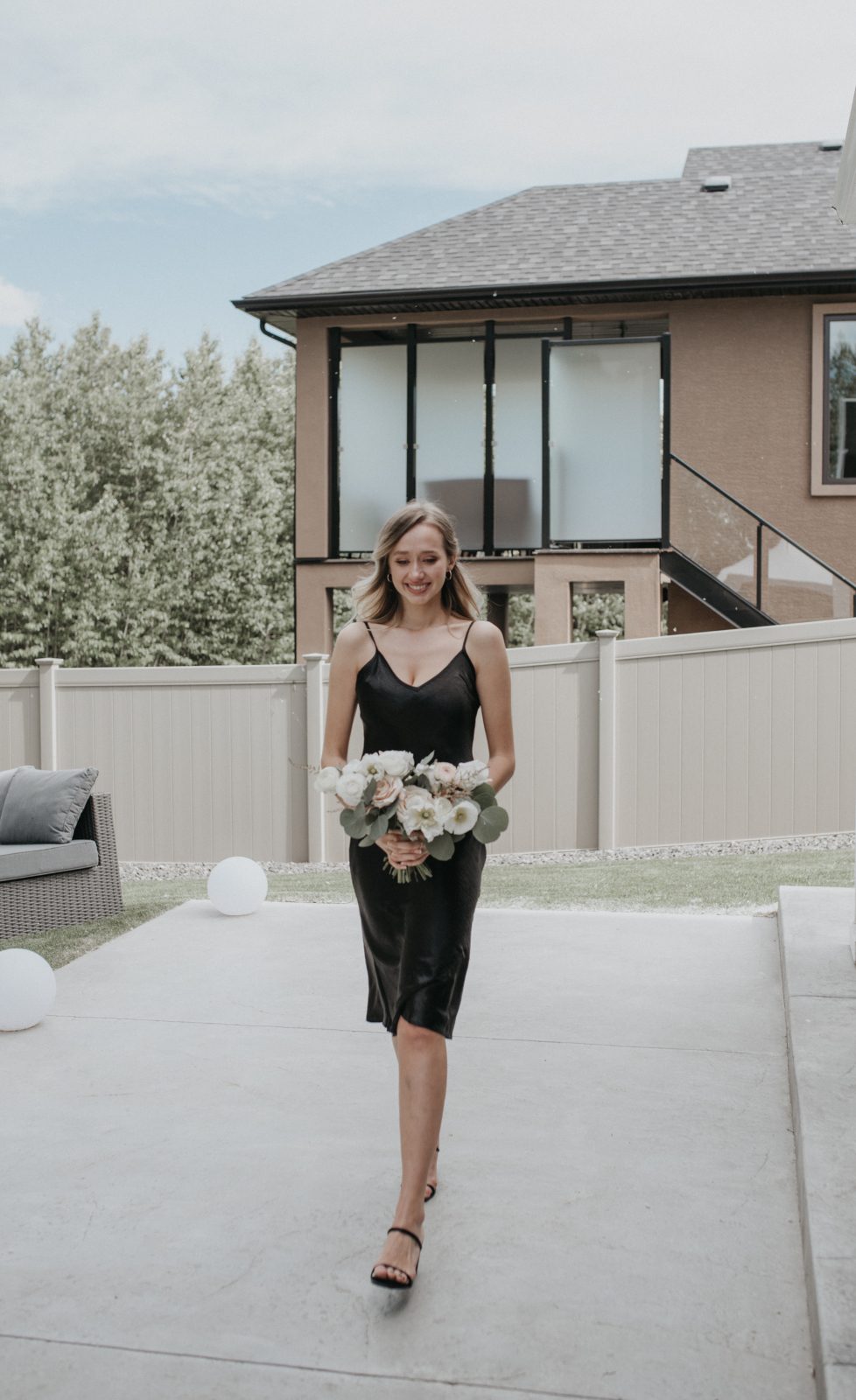 The bridesmaid walks with a soft smile, holding a round bouquet of white and blush flowers with fresh greenery, wearing a sleek black satin gown.