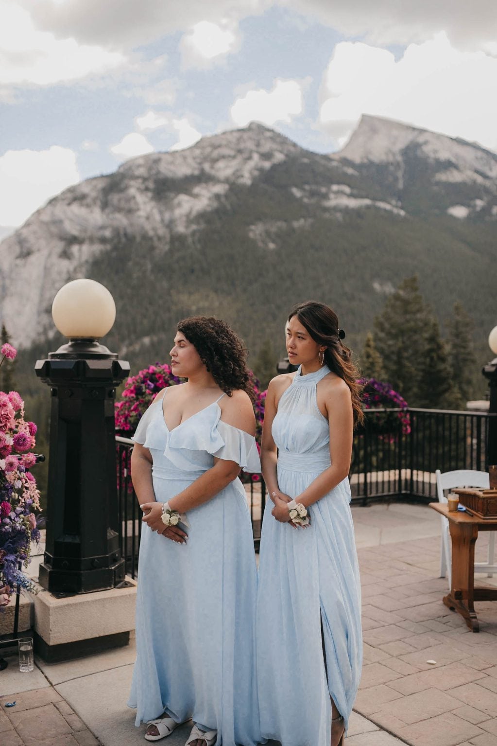 Two bridesmaid in flowing light blue gowns watch the ceremony with serene expressions as mountains rise behind them.