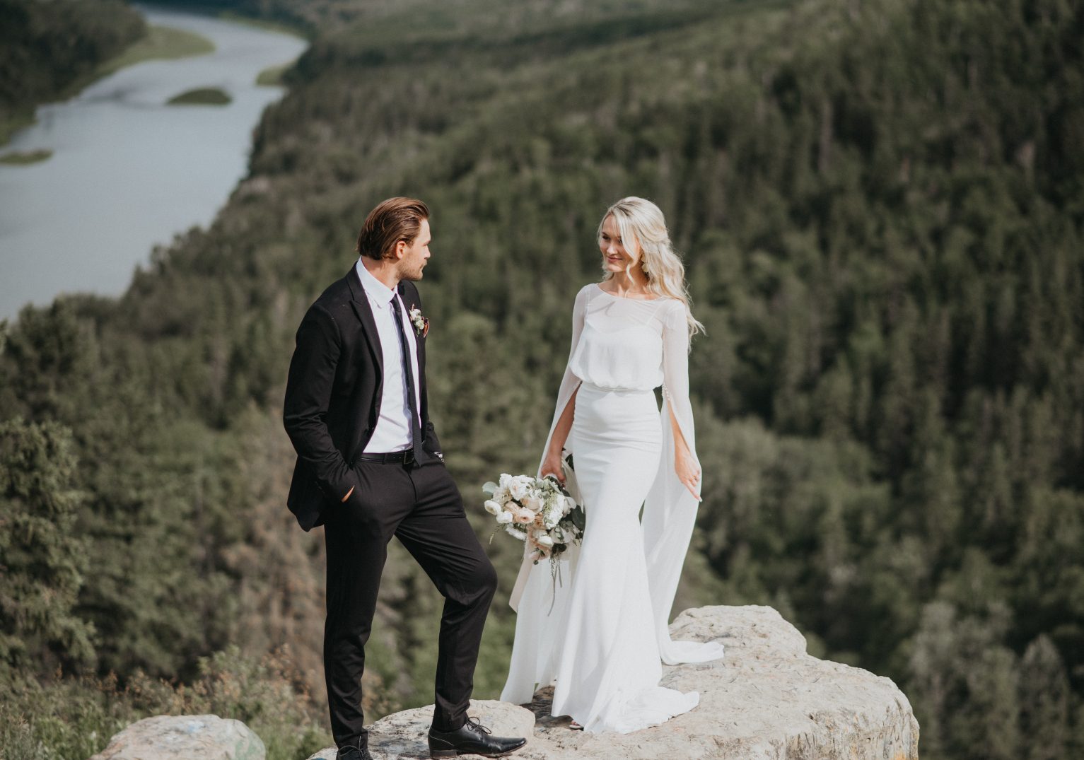 The bride and groom stand atop a rocky cliff overlooking a lush valley, exchanging a loving glance in their wedding attire.