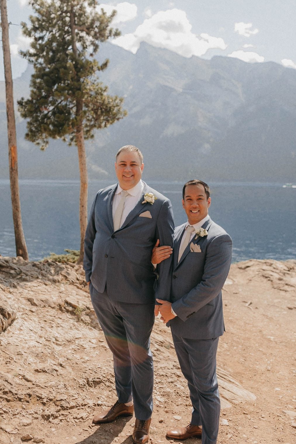 Two grooms in blue suits smile together besides a crystal blue lake with mountains rising in the background.