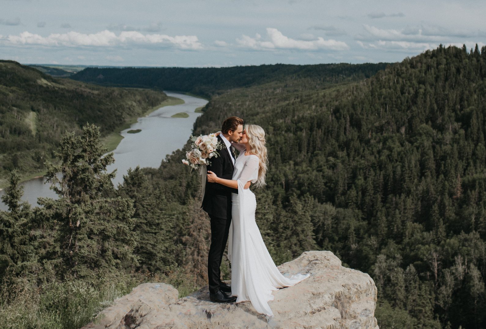 The bride and groom share a kiss on a scenic cliff, surrounded by forested hills and a winding river below.