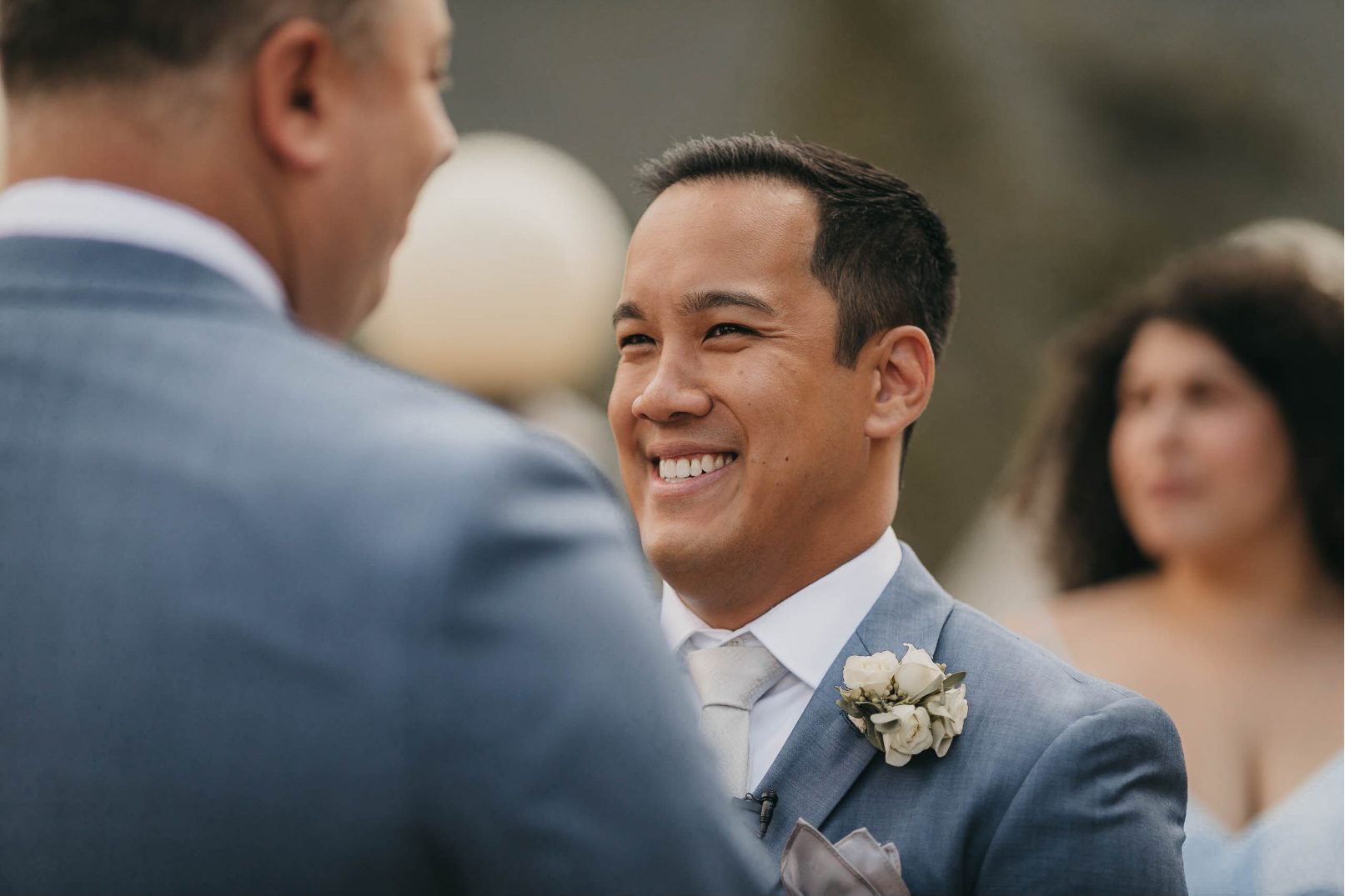 Close up of one groom smiling with emotion during the vows, a white spray rose boutonniere on his blue suit.