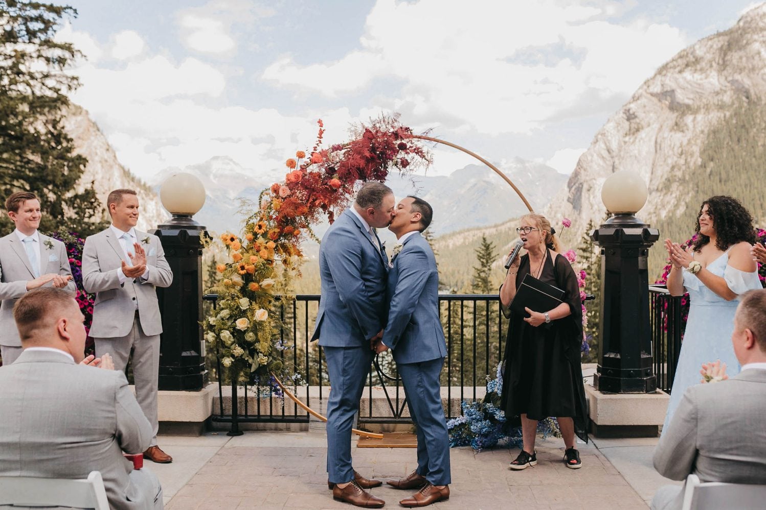 Two grooms share their first kiss as newlyweds beneath a vibrant rainbow floral arch on the terrace at Banff Springs