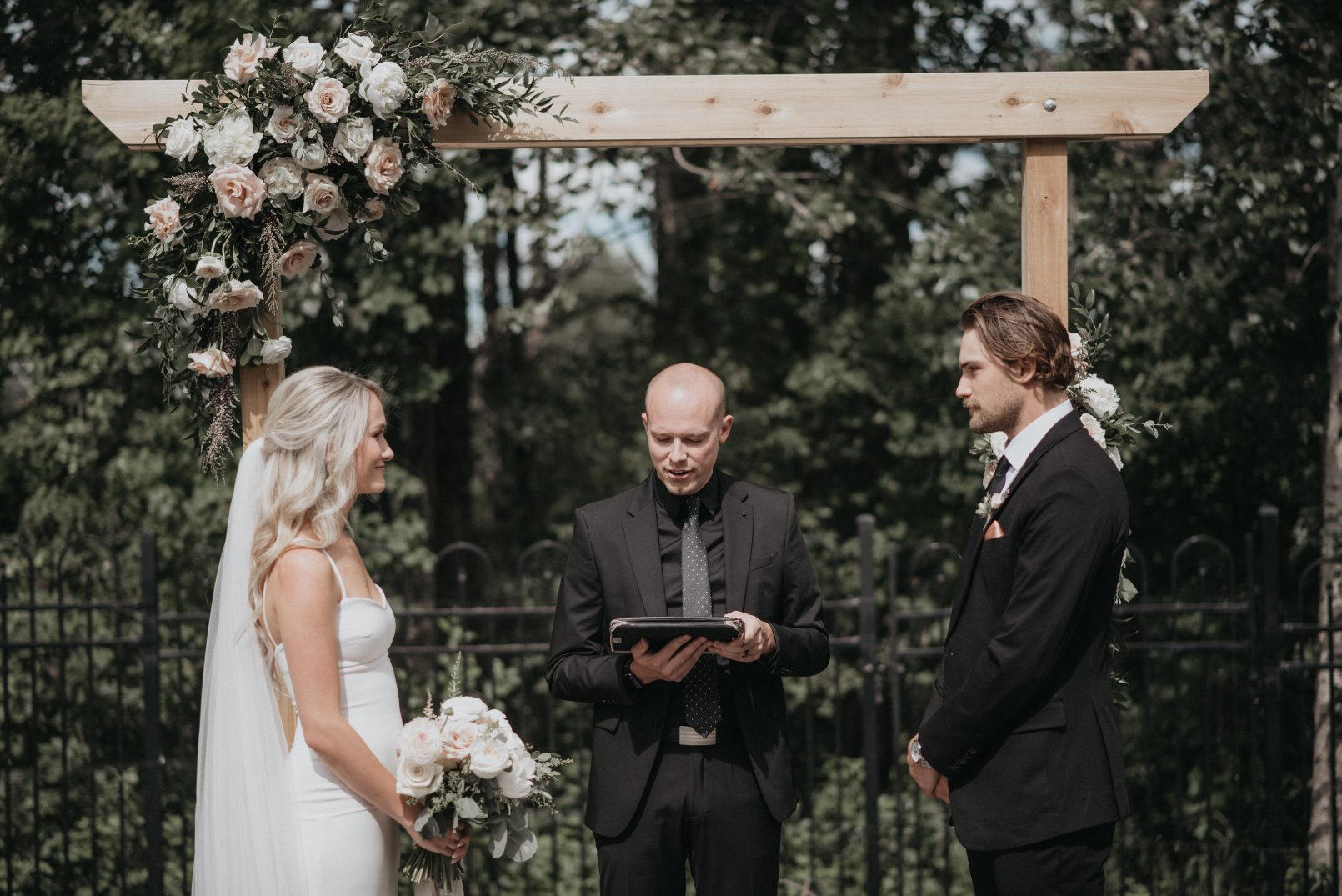 The couple stands before an officiant under a wooden floral arch, smiling as they exchange vows during their outdoor ceremony.