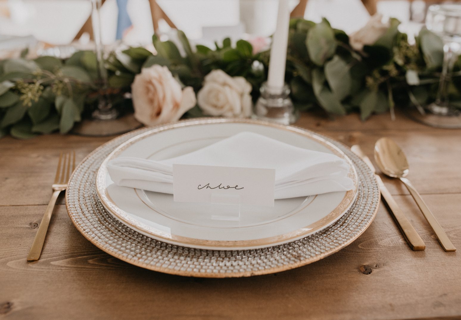 A detailed table setting featuring gold cutlery, a textured glass charger plate, elegant white china and a place card labelled "Chloe."