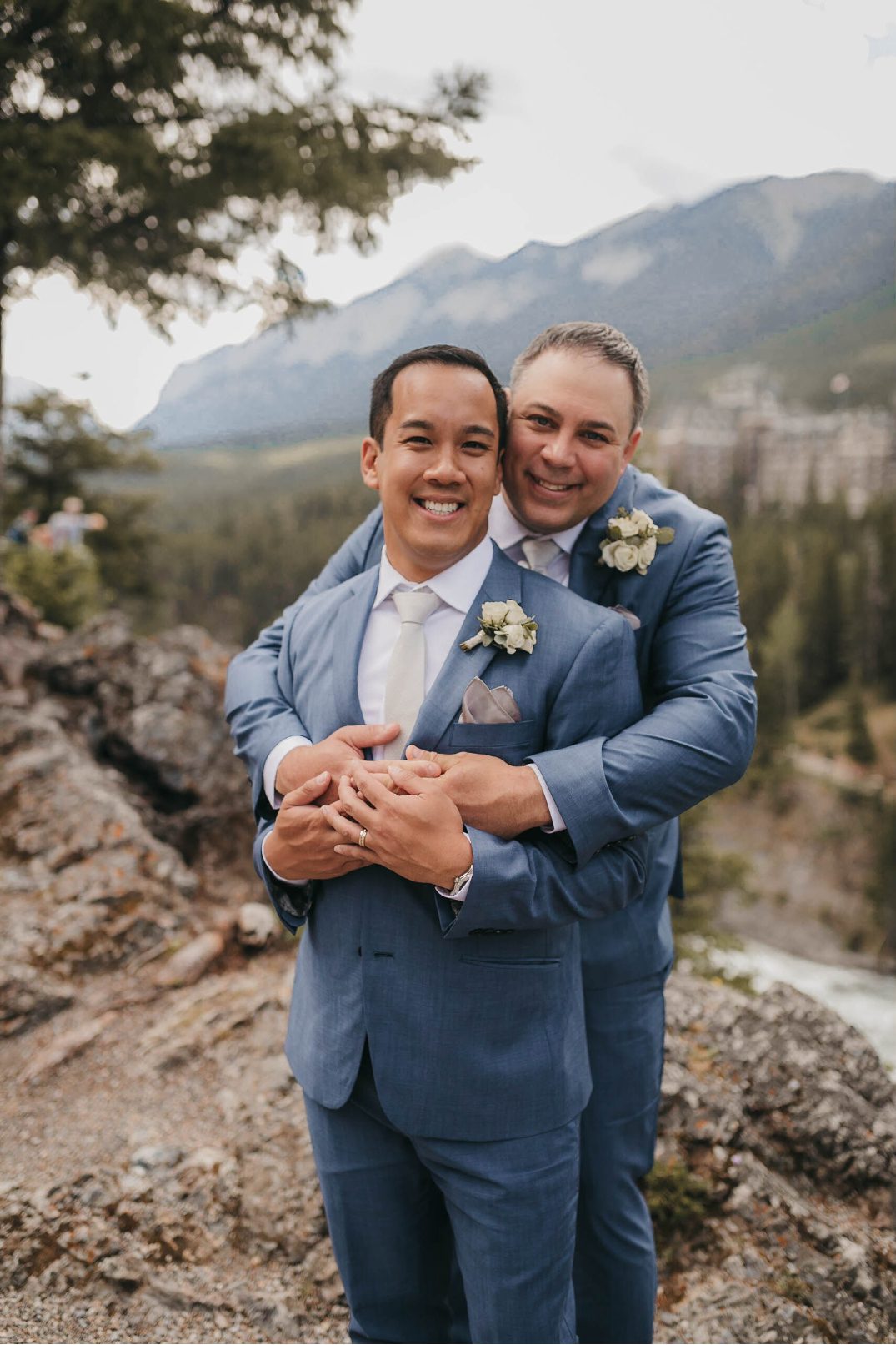 One groom embraces the other from behind as they smile warmly, with forested mountains in the distance.