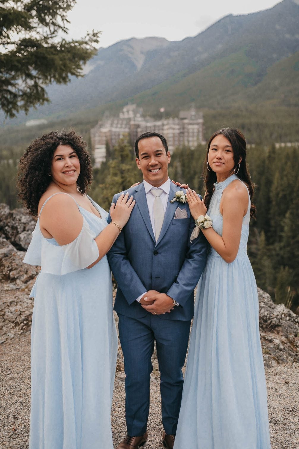 One groom stand smiing between two bridesmaids in powder blue dresses with the hotel visible behind them.