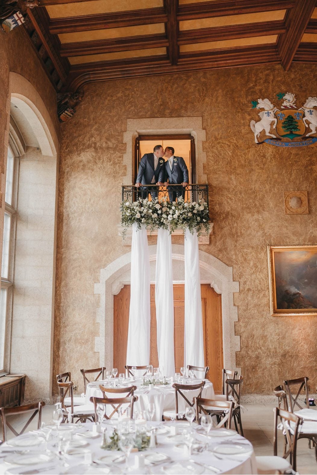 Two grooms kiss while standing on an indoor balcony decorated with white florals and soft draping fabric.