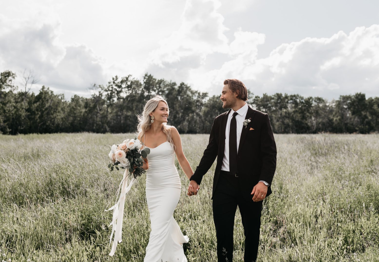 The bride and groom walk hand in hand through a grassy field, both smiling as the bride holds a lush bouquet of white and blush blooms.