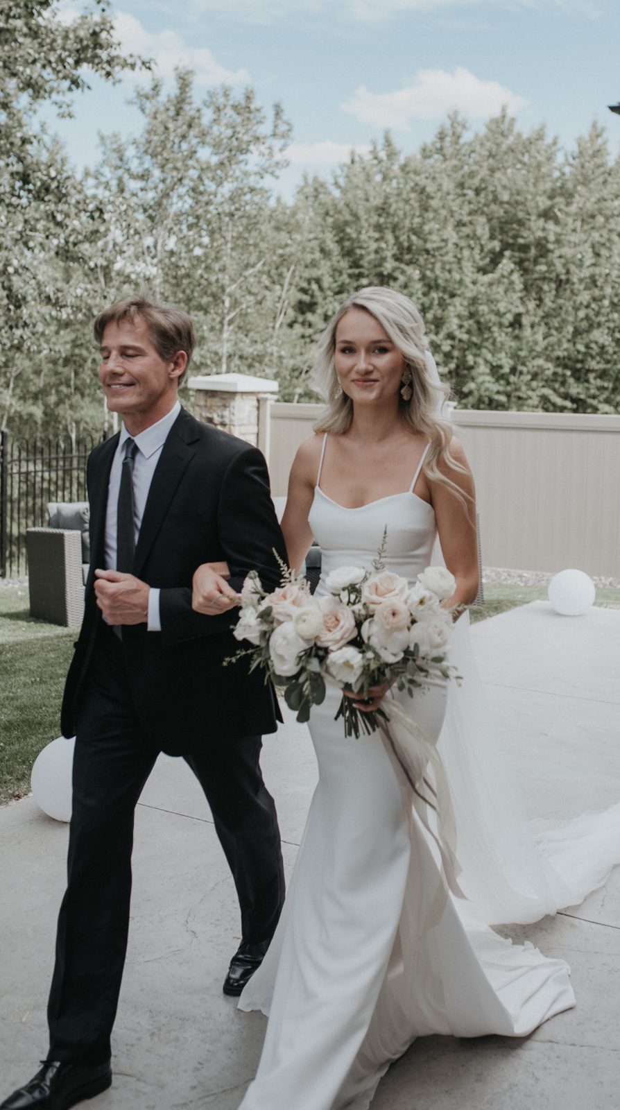 The bride walks arm in arm with her father towards the ceremony, holding a romantic bouquet of blush and white roses with trailing silk ivory ribbons.