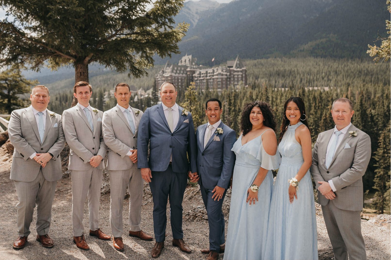 The wedding party poses in front of the Fairmont Banff Springs Hotel, dressed in soft blue and grey tones.