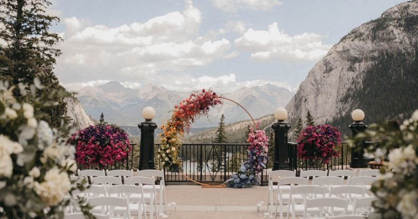 A vibrant floral arch in full rainbow colours, celebrating love, stands on a terrace, framed by mountains and a cloudy blue sky.