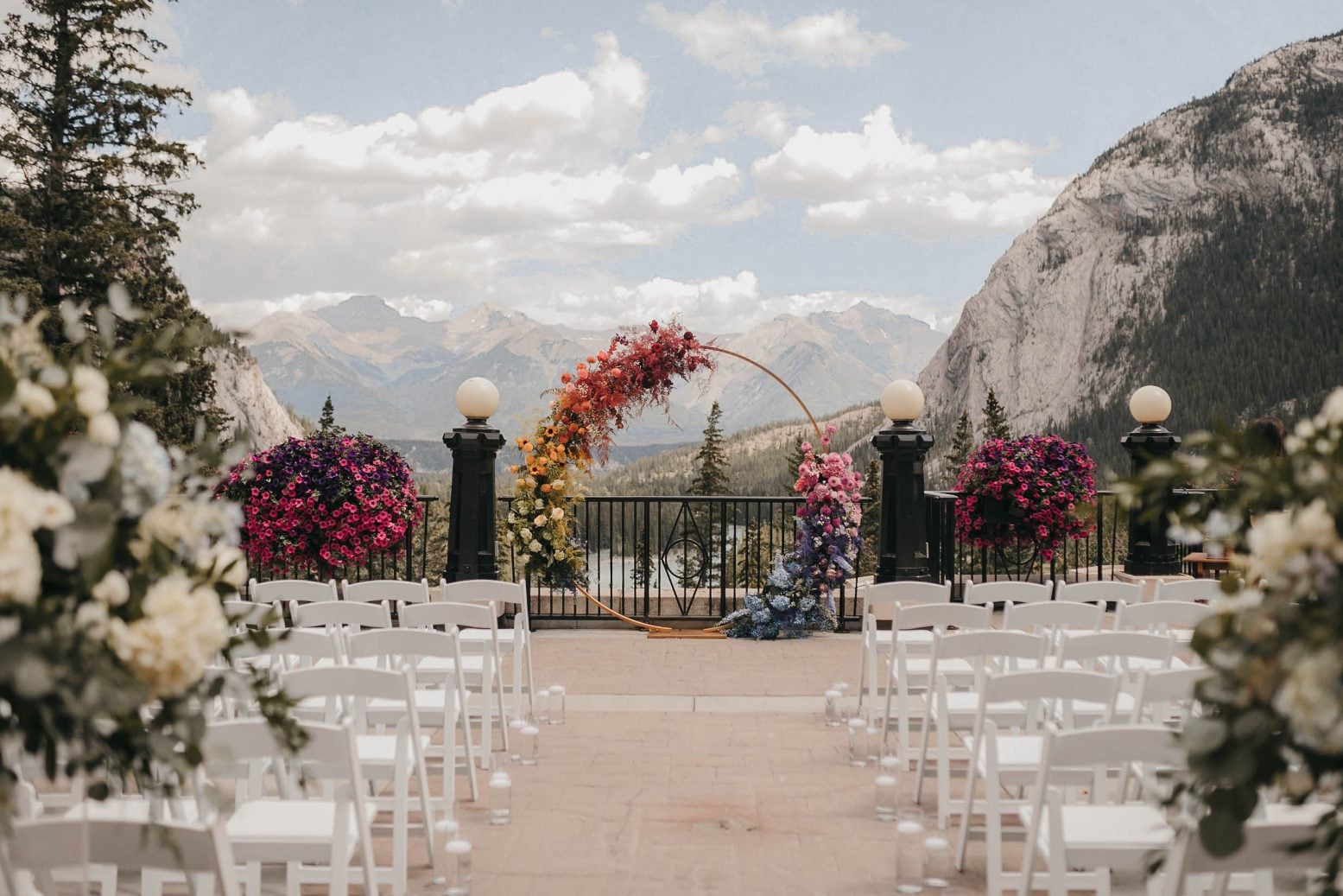 A vibrant floral arch in full rainbow colour stands on a terrace for the ceremony, framed by mountains and a cloudy blue sky.