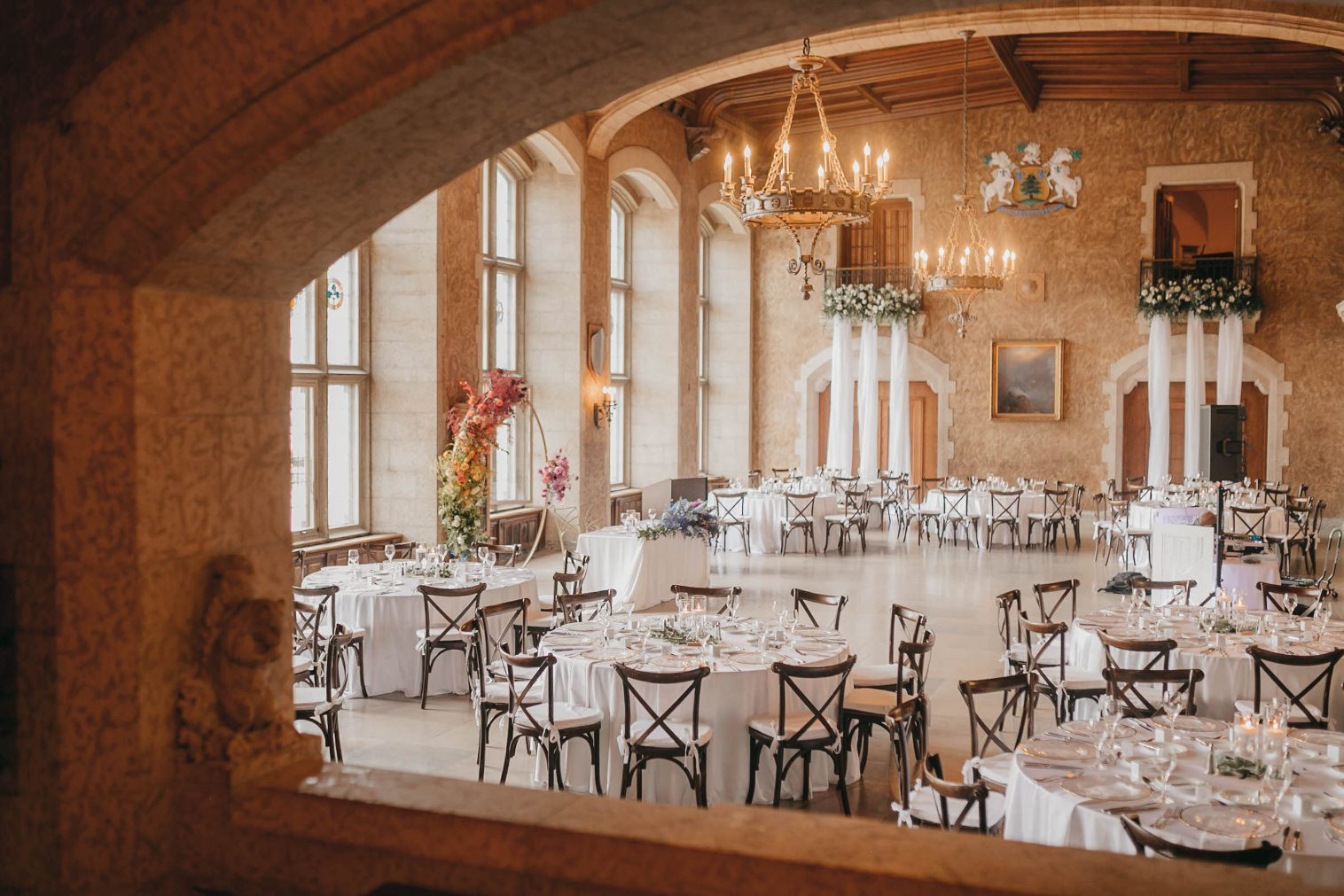 A sweeping view of the reception hall featuring ornate chandeliers, tall windows, and the rainbow floral arch repurposed inside.