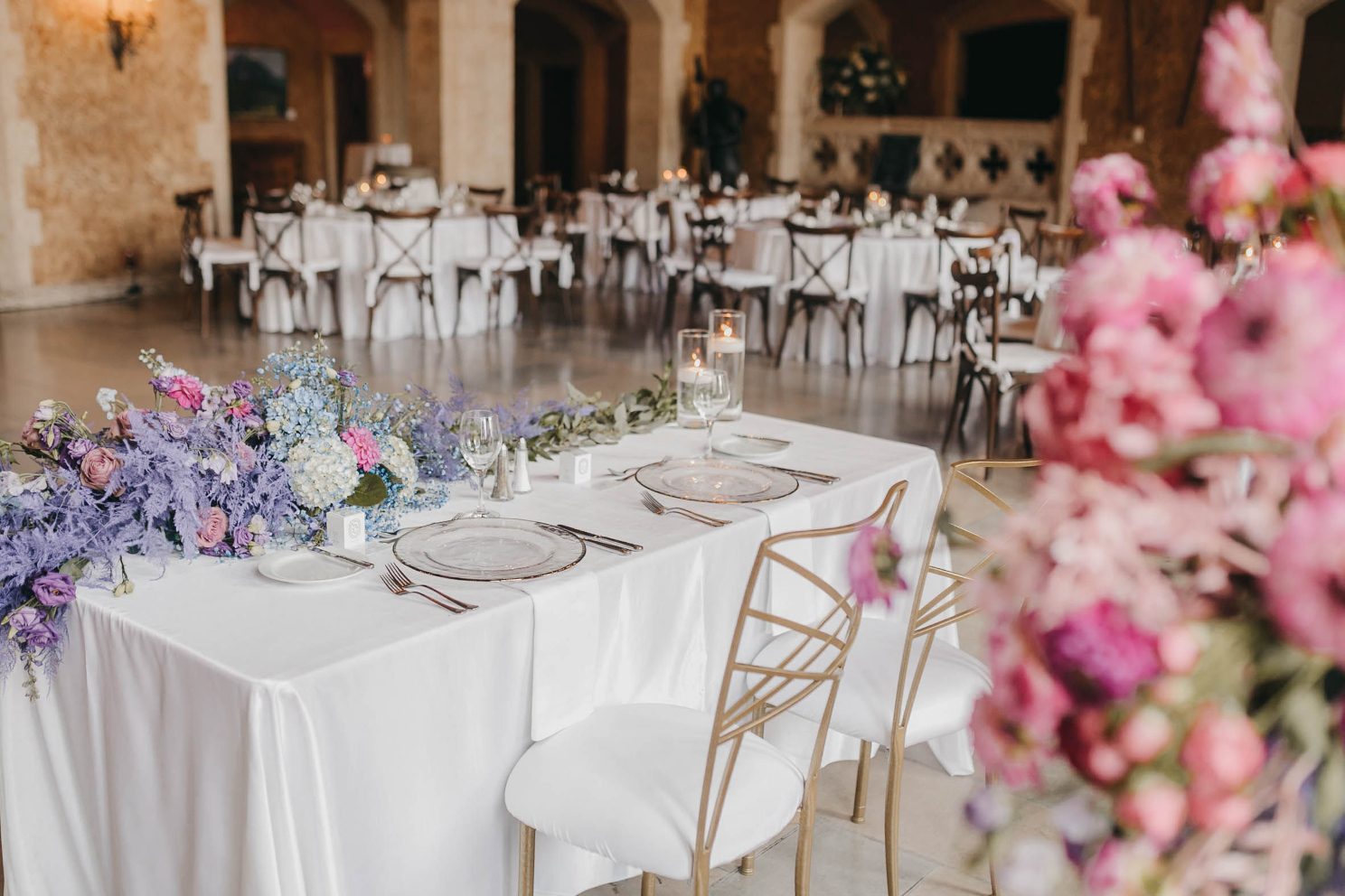 A sweetheart table adorned with purple and blue florals, set with modern glassware and gold chairs.