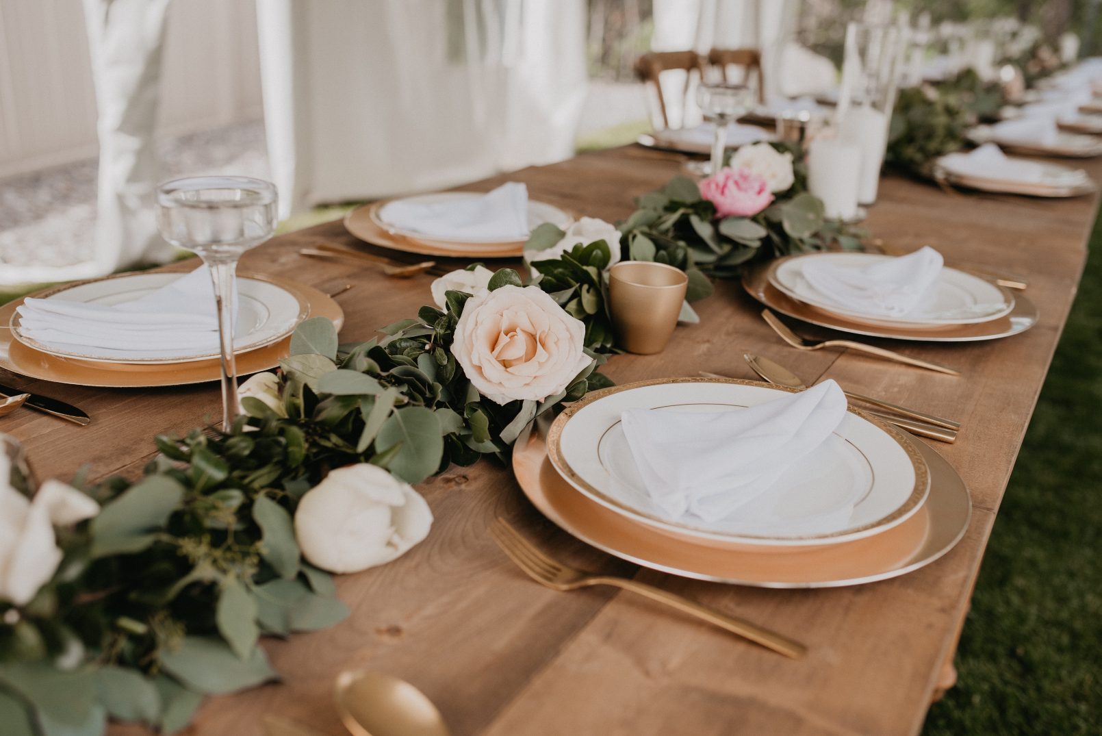 A long wooden reception table decorated with a lush green garland of eucalyptus, blush roses, white napkins and gold accented tableware.