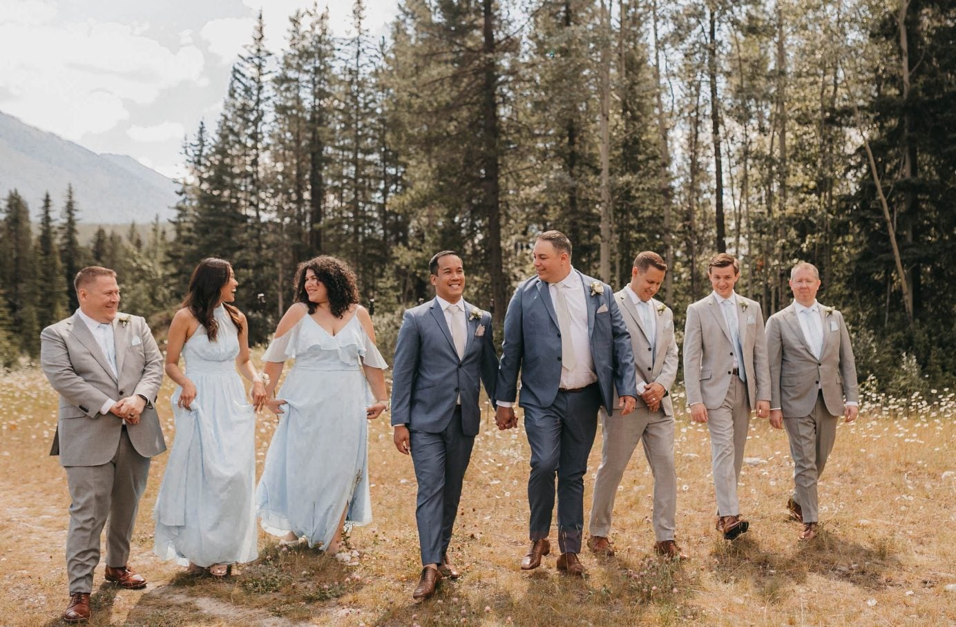 The grooms walk hand in hand with their wedding party through a meadow surrounded by forest.