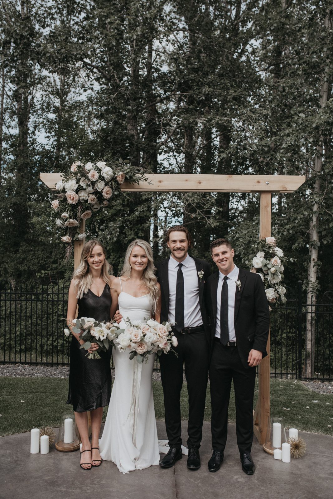 The bride and groom stand under a wooden ceremony arch adorned with soft blush and white florals, flanked by a bridesmaid and groomsman in black attire.