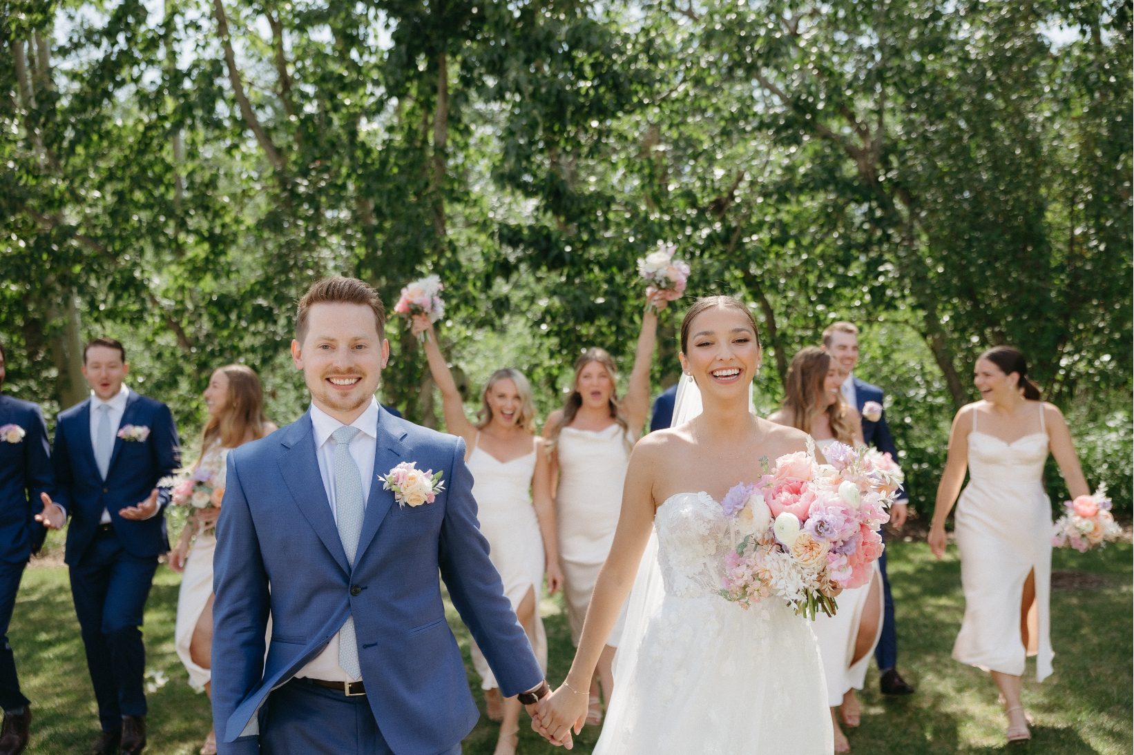 The bride and groom walk hand in hand, beaming with joy, surrounded by their lively wedding party dressed in coordinating blue suits and ivory dresses with lush greenery as the backdrop.
