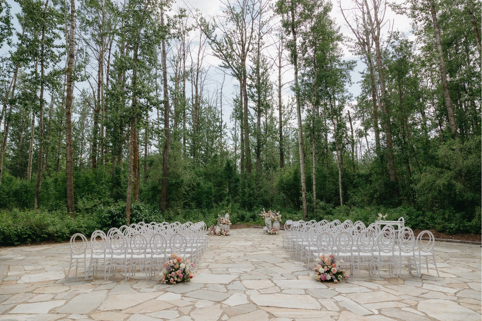 The ceremony set up at Sparrow Lane with rows of white chairs, two pastel floral arrangements marking the aisle entrance, and a cluster of arrangements in urns and on pillars framing the alter.