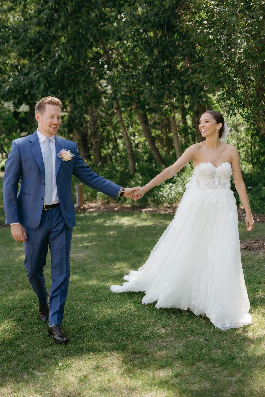 The newlyweds smile at each other while walking hand in hand on a sun dappled lawn, the brides floral lace gown trailing behind her.