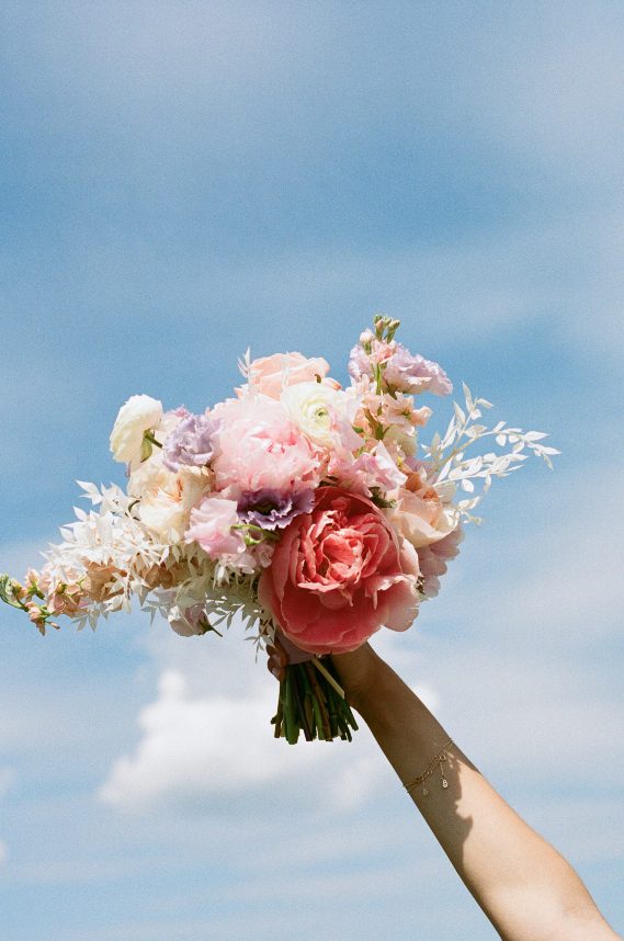 A close up of the bride's bouquet held high against a bright blue sky, featuring pink peonies, blush roses, white ranunculus and soft lavender blooms.