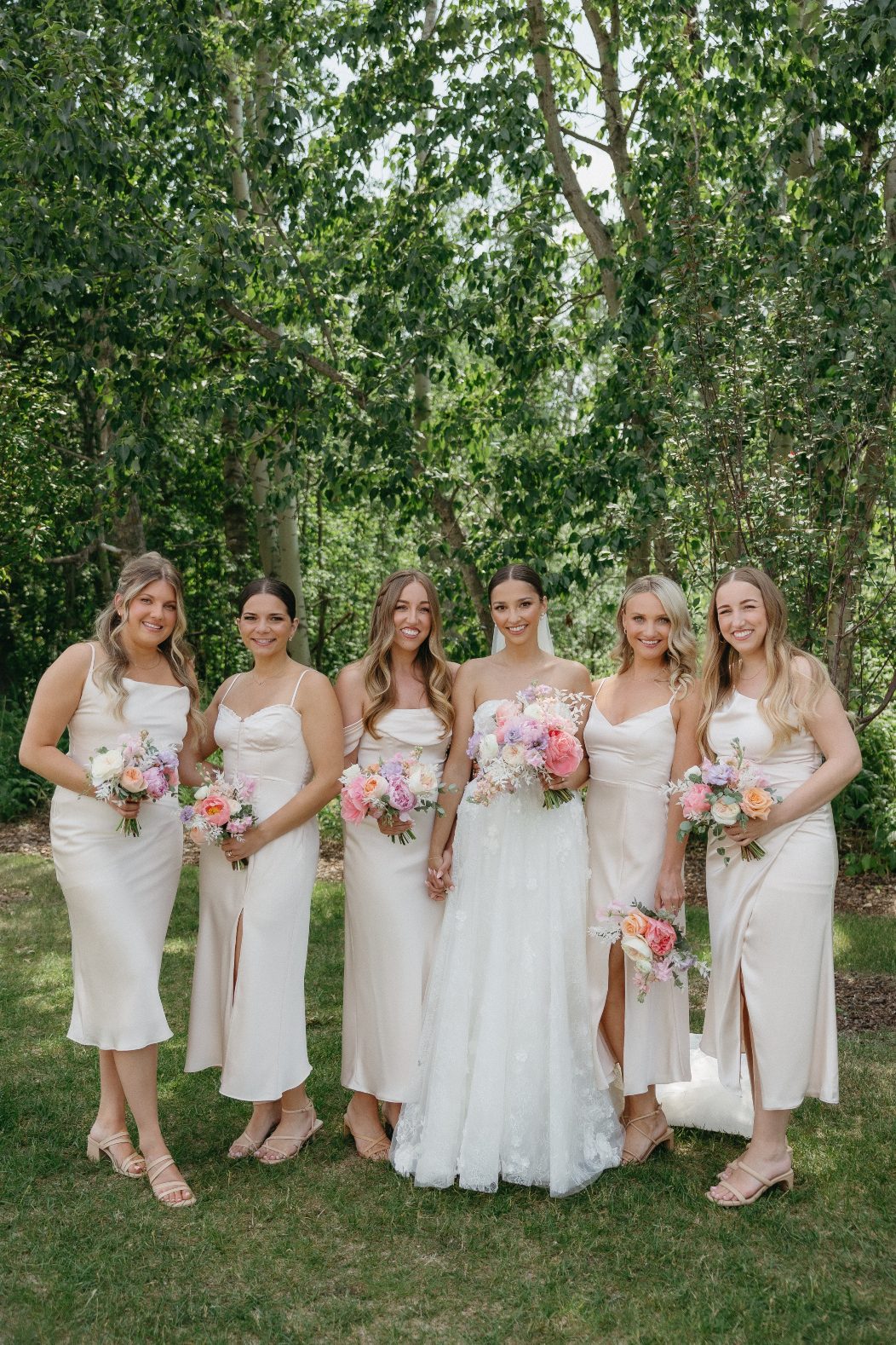 The bride poses with her five bridesmaids, all in matching champagne-toned satin dresses, each holding bouquets in soft pastel tones