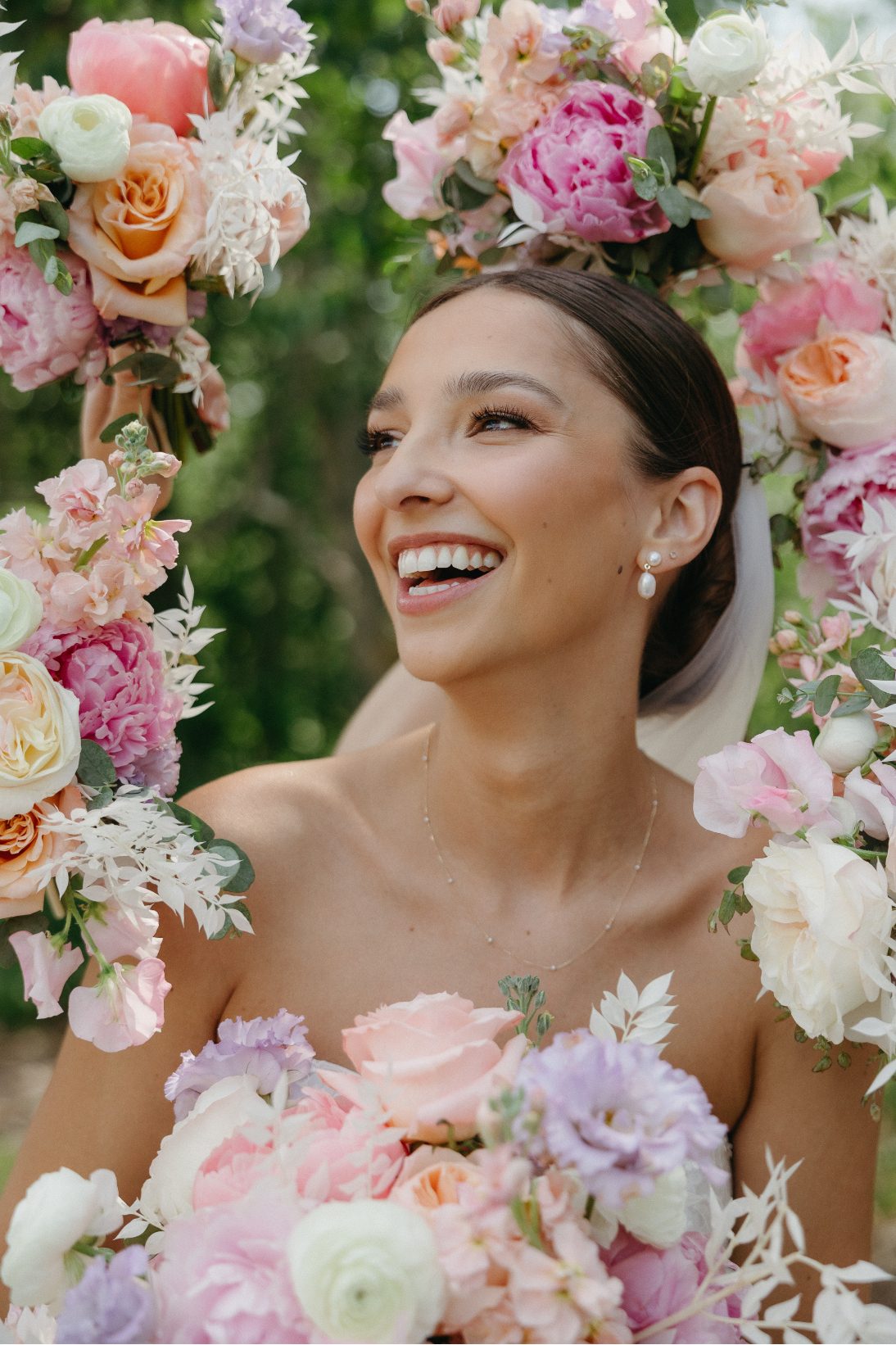 A joyful close up of the bride, framed by vibrant wedding florals in shades of peach, blush, lavender and ivory as she smiles brightly.