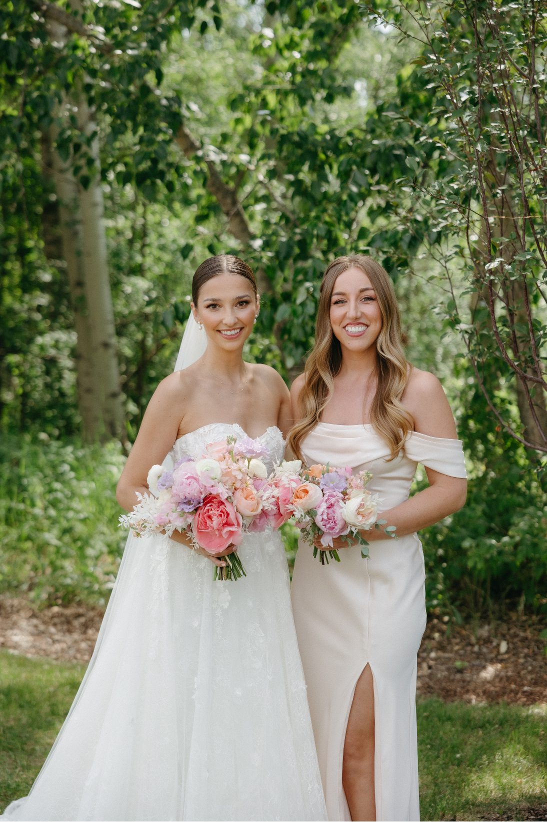 A portrait of the bride and a bridesmaid standing side by side, smiling softly at the camera with matching blush toned bouquets and dresses.