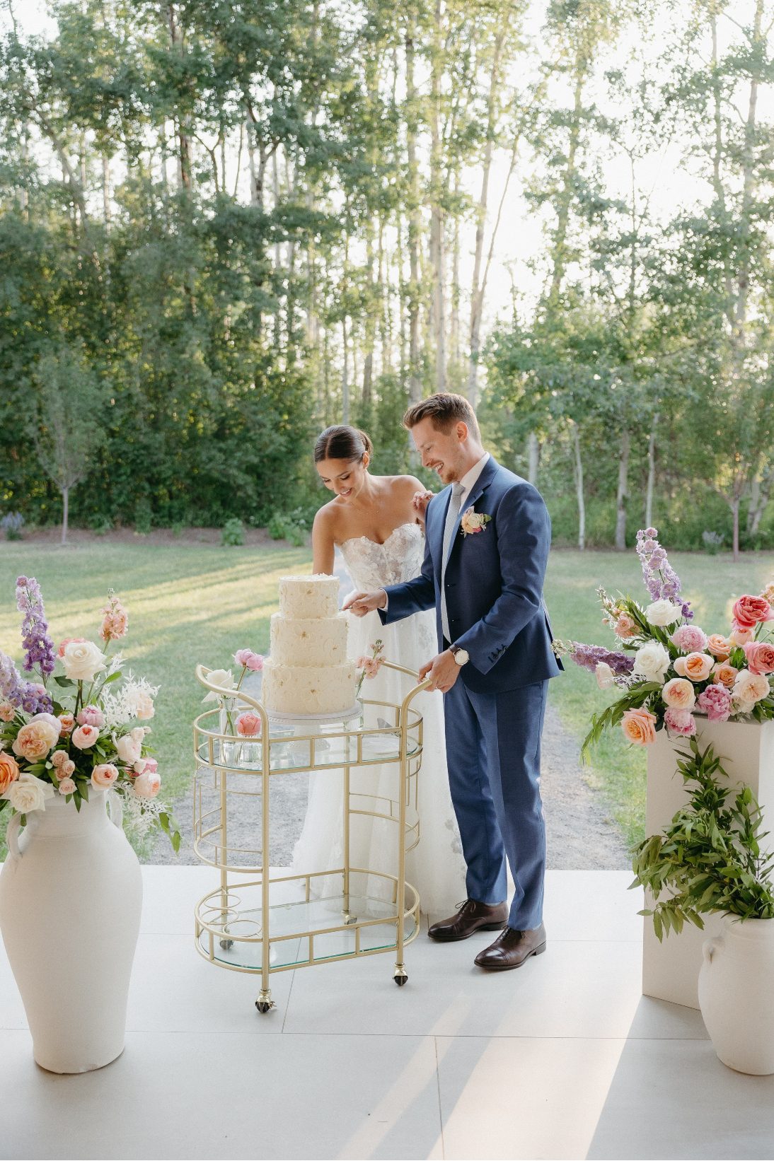 The bride and groom stand together in the sunshine cutting their cake together.